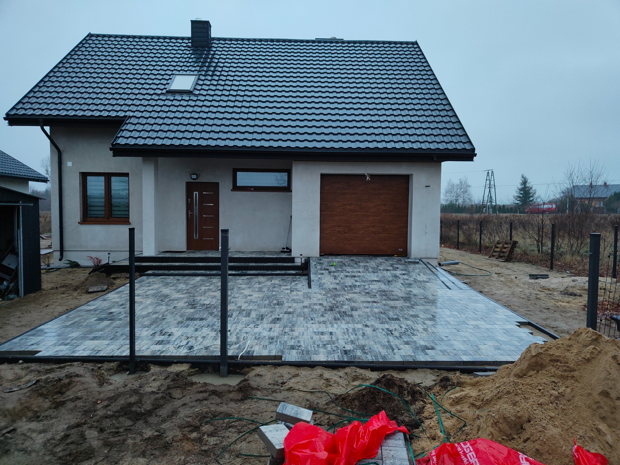 Nowoczesny dom jednorodzinny z częściowo ułożoną, szaro-białą kostką brukową na podjeździe. W tle widoczne drzewa i inne budynki, a w pierwszym planie stos kostki zabezpieczony czerwoną folią.