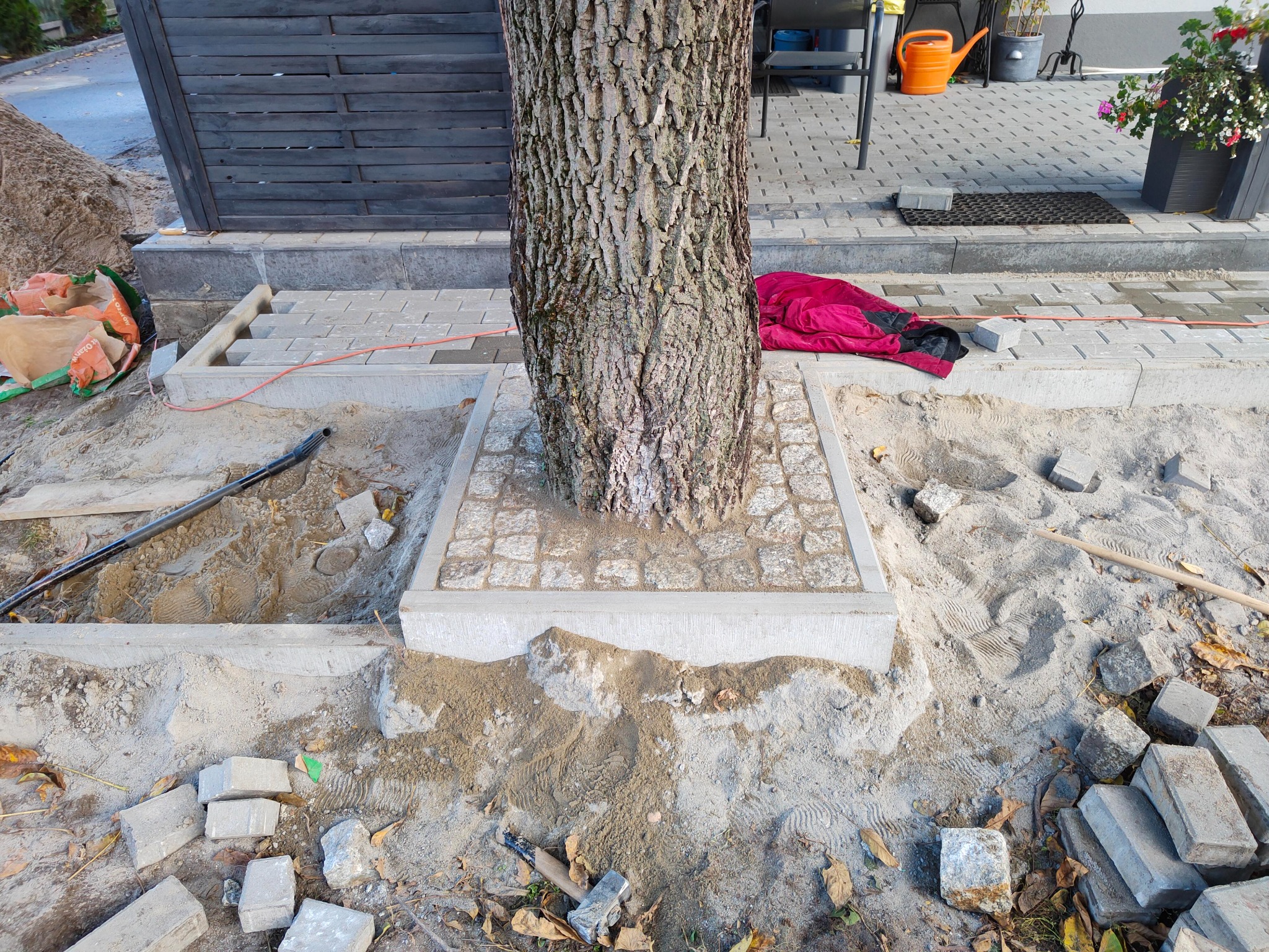 Niedokończona nawierzchnia z kostki wokół drzewa, widoczne narzędzia, piasek i przygotowane obrzeża, w tle fragment domu z szarą elewacją.