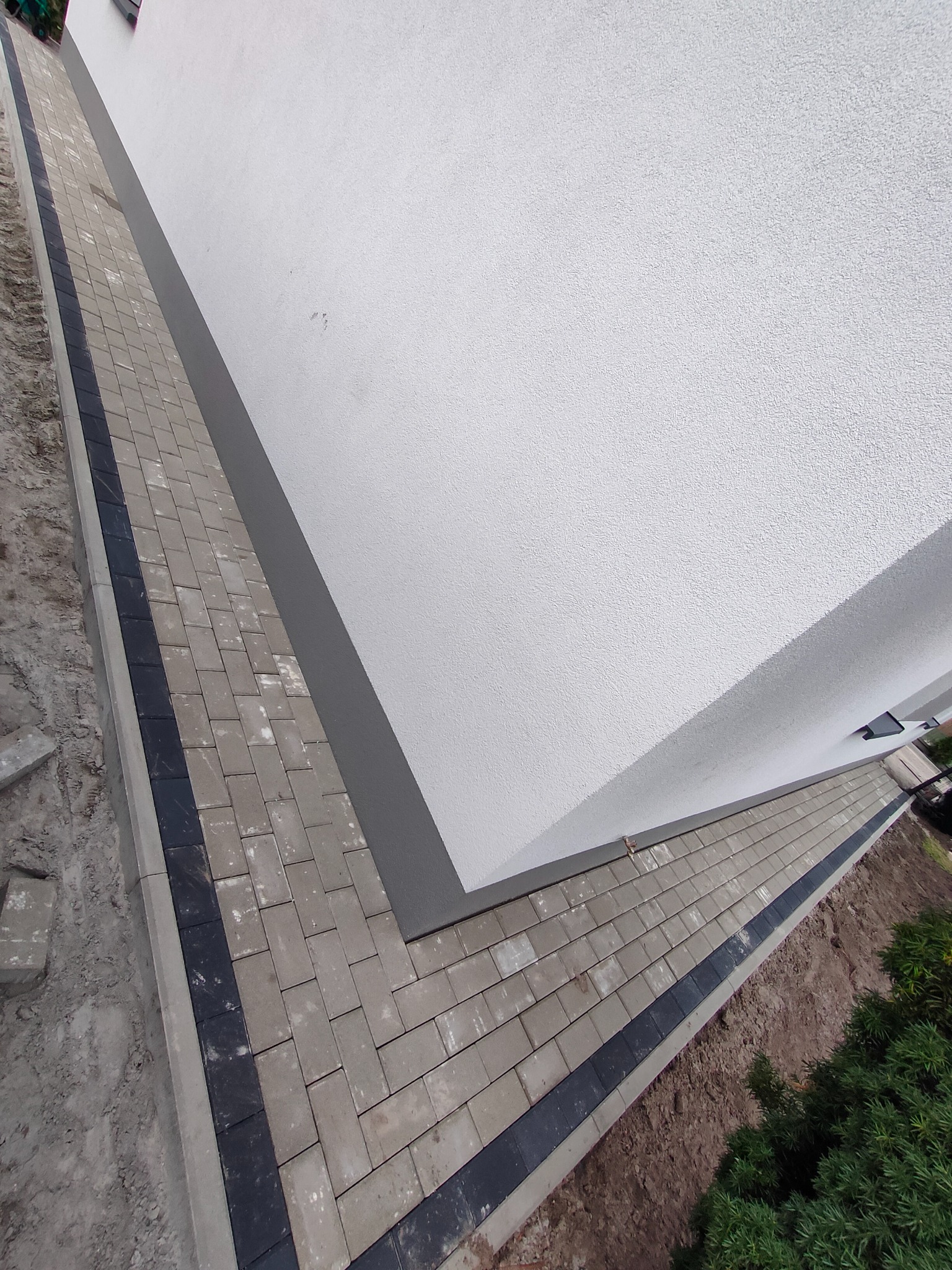 Świeżo ułożony chodnik z jasnoszarej kostki brukowej z czarnym obramowaniem, przylegający do białej ściany budynku, widoczny fragment ziemi i zieleni.