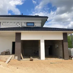 Niedokończony dom jednorodzinny z widocznymi pustakami, czarnym dachem, garażem w stanie surowym, piaskiem na podwórku, paletą z pojemnikiem na wodę i rowerem w garażu.