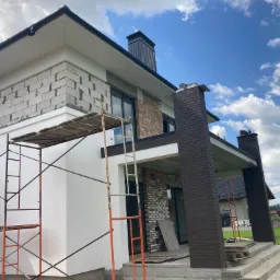 Dom jednorodzinny w trakcie budowy: widoczne rusztowania, ściany z bloczków, fragment elewacji z cegły klinkierowej i biały tynk, kominy z blachy na dachu.