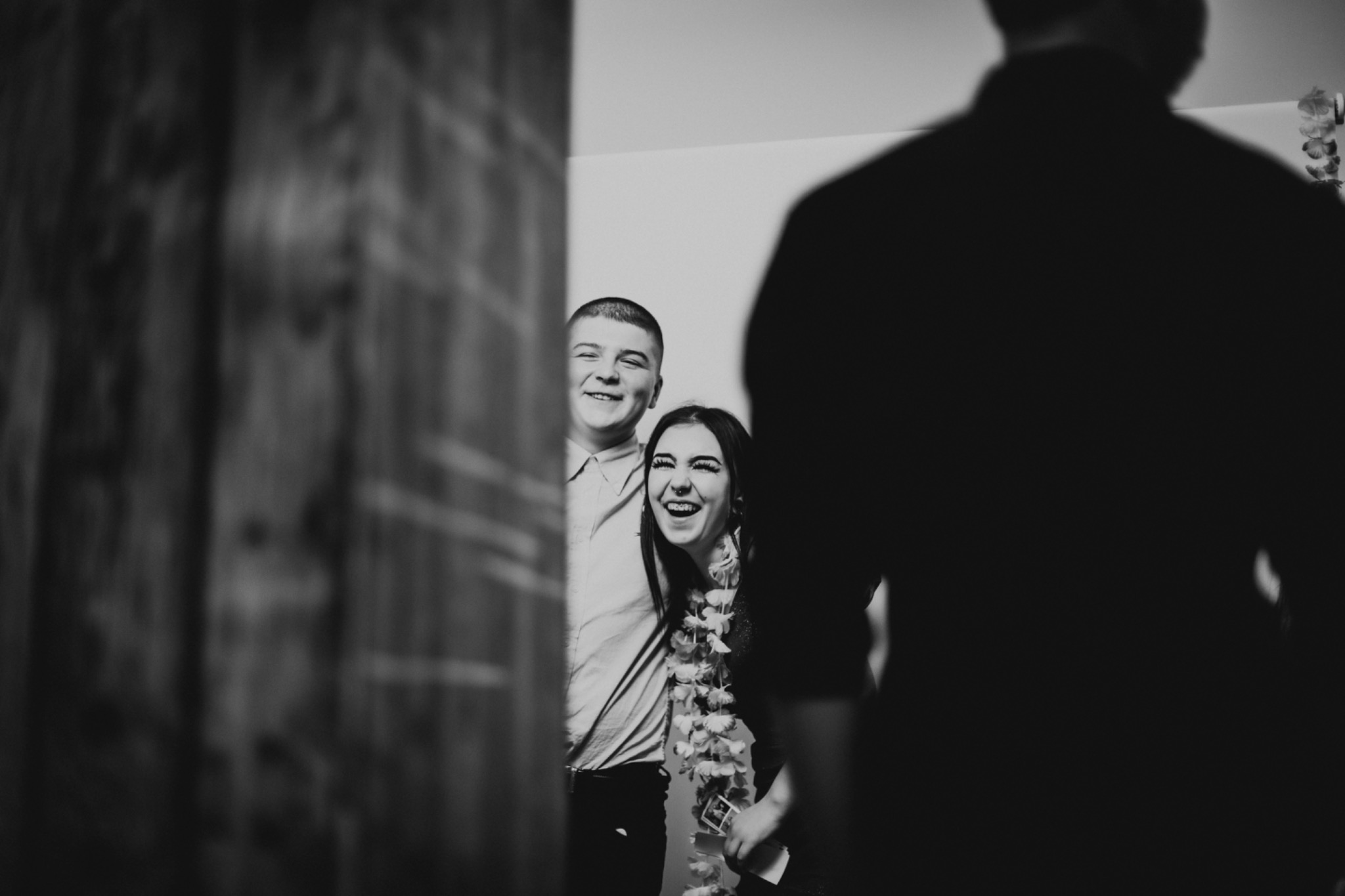 Czarno-białe zdjęcie z fotobudki: uśmiechnięta para w hawajskich girlandach, z widocznym fragmentem zdjęć z sesji, w tle zamazana sylwetka osoby.