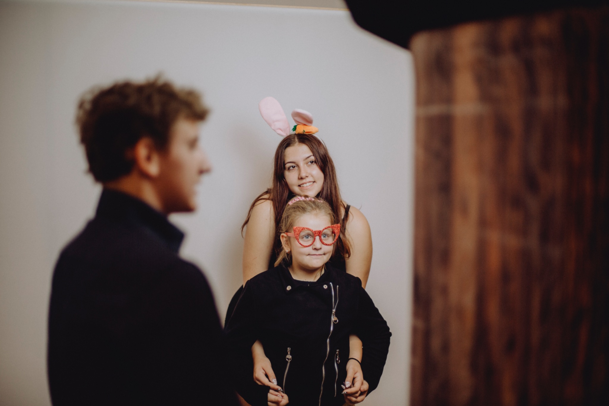 Trzy osoby pozują do zdjęcia w fotobudce: dziewczynka w okularach w kształcie serc i czarnej kurtce, za nią nastolatka w uszach królika i chłopiec z lewej strony kadru.