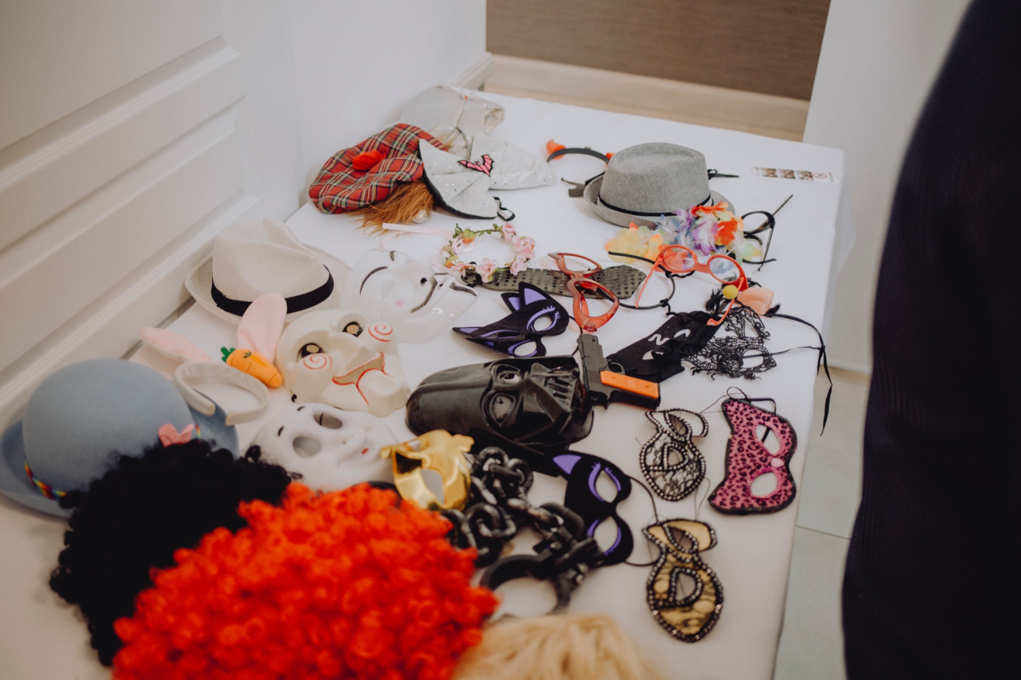 Różnorodność rekwizytów do fotobudki: maski karnawałowe, kolorowe peruki, kapelusze, okulary leżące na białym stole.