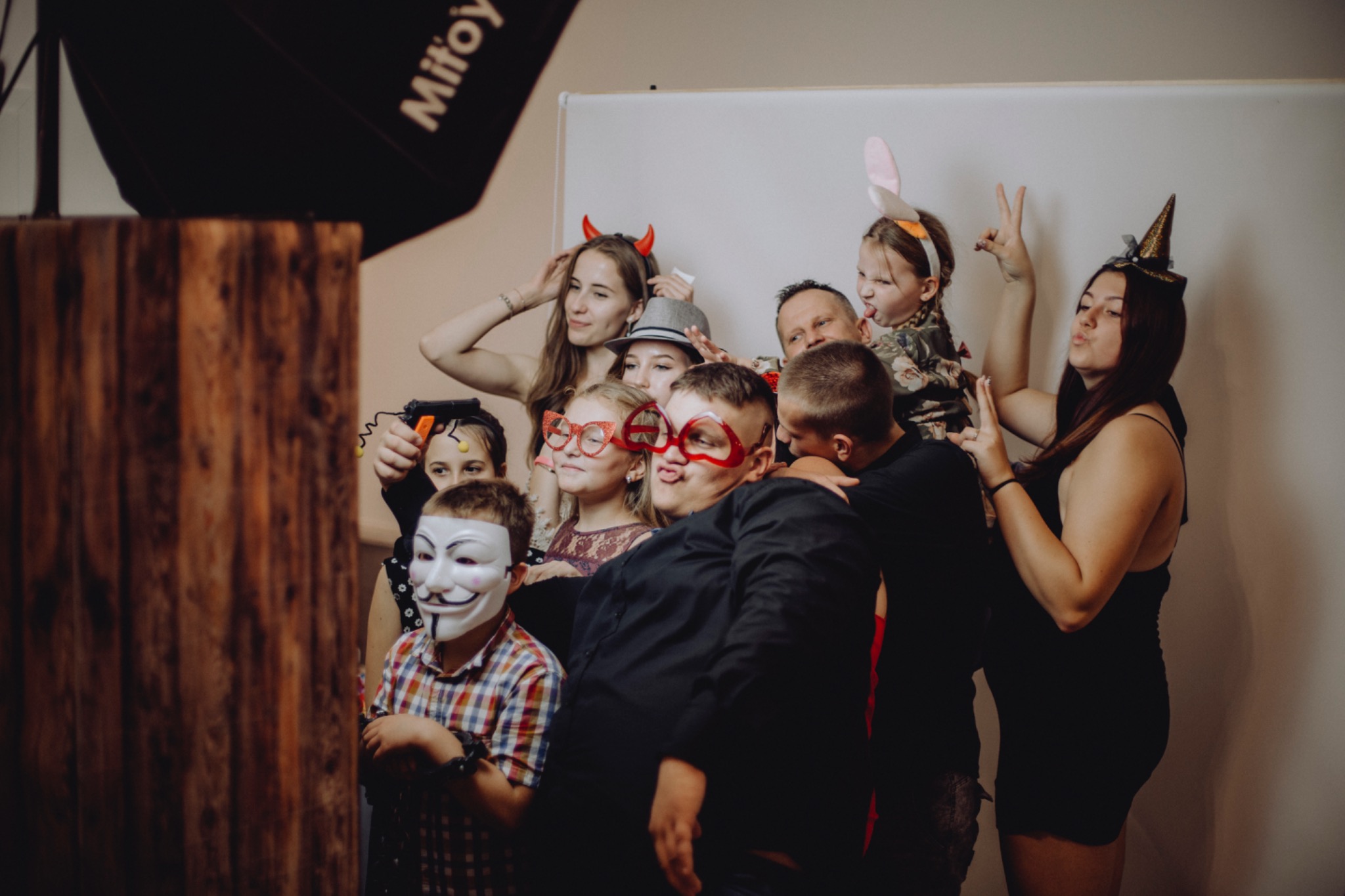 Grupa osób w przebraniach pozuje do zdjęcia w fotobudce z rekwizytami, w tym maskami, okularami i rożkami diabła.