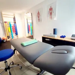 Wnętrze gabinetu rehabilitacyjnego z szarym stołem do masażu z zielonym wałkiem, drabinkami gimnastycznymi, piłką do ćwiczeń i sprzętem do fizykoterapii.