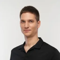 Portret mężczyzny w czarnej koszulce polo na jasnym tle, z krótkimi, ciemnymi włosami i lekkim uśmiechem, prezentujący profesjonalny wizerunek.