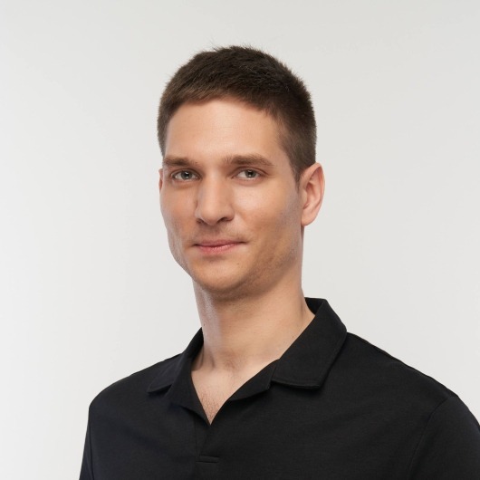 Portret mężczyzny w czarnej koszulce polo na jasnym tle, z krótkimi, ciemnymi włosami i lekkim uśmiechem, prezentujący profesjonalny wizerunek.