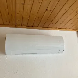 Biała jednostka klimatyzacyjna marki Gree zamontowana na ścianie z dekoracyjną okładziną imitującą kamień, widoczny drewniany sufit z boazerią.