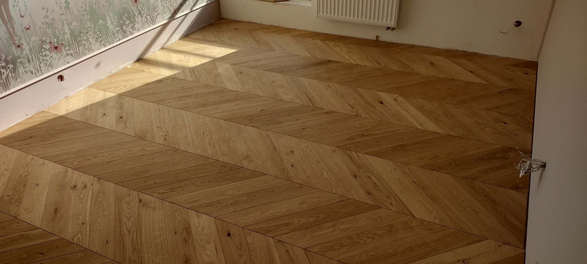 Ułożony parkiet w jodełkę francuską, widoczne niedokończone prace wykończeniowe przy ścianie i wystające kable.