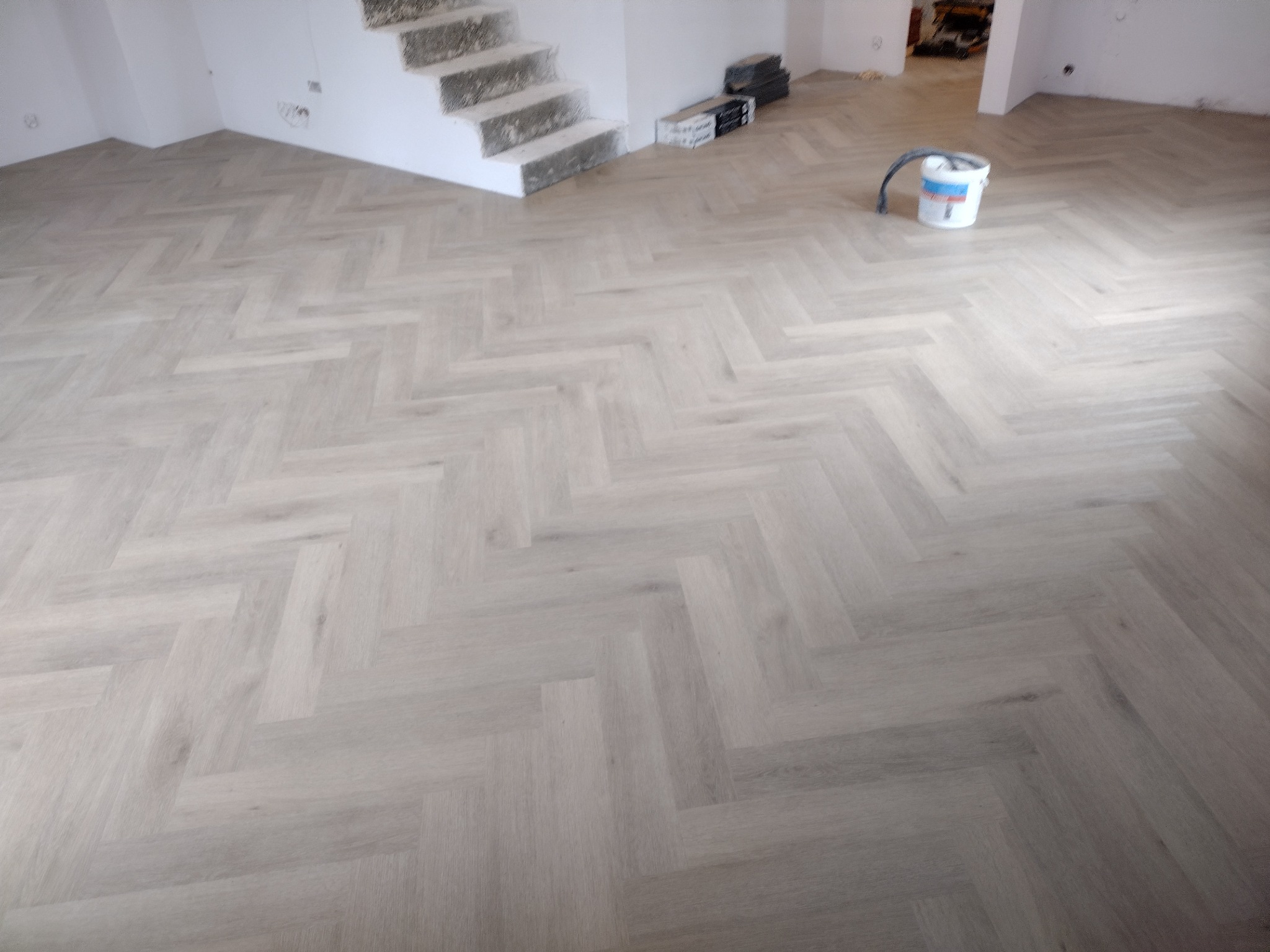 Ułożony jasnoszary parkiet w jodełkę francuską w przestronnym pomieszczeniu, widoczne schody w tle i wiadro z klejem.