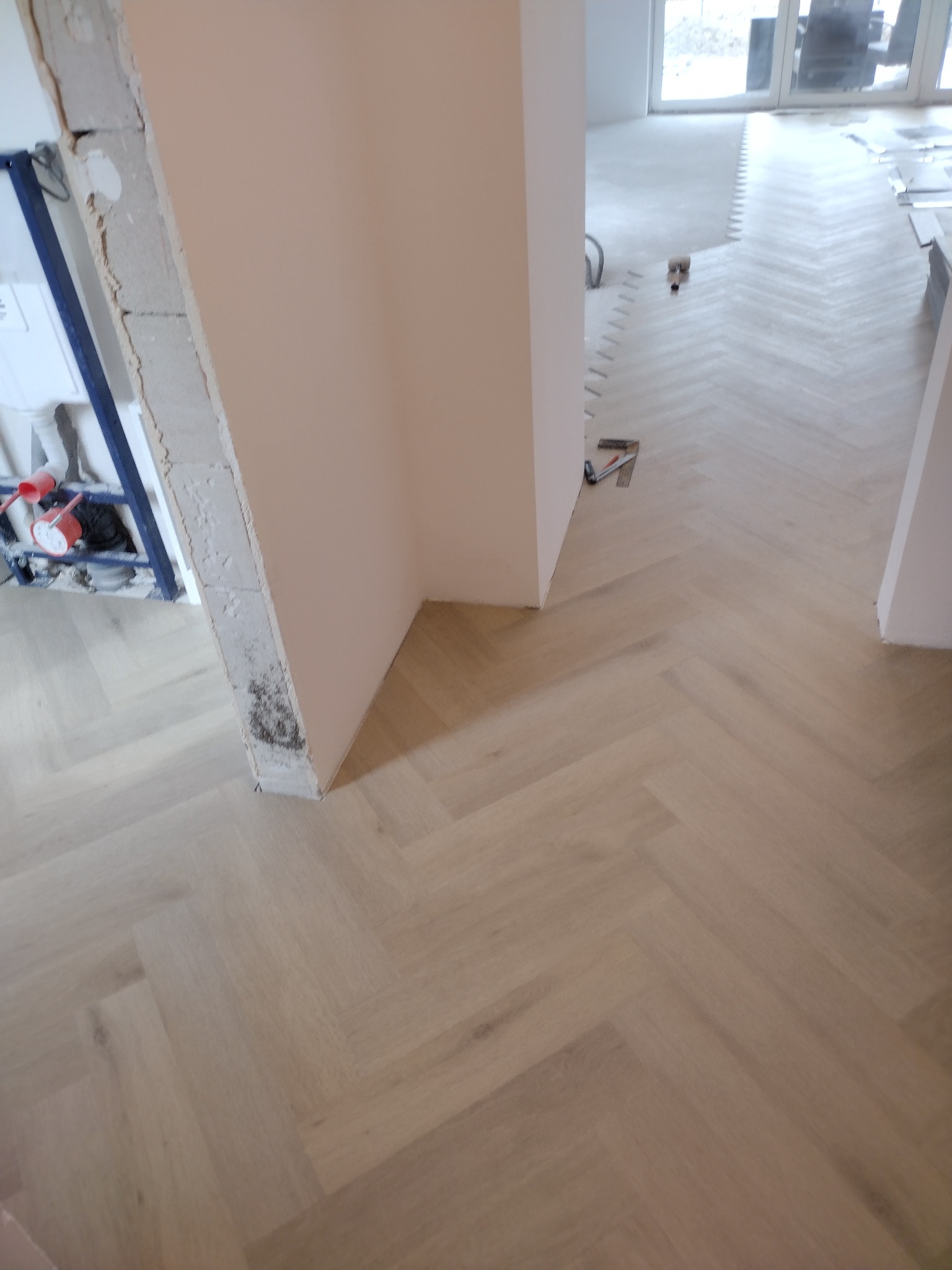 Jasny parkiet ułożony we wzór jodełki francuskiej w trakcie montażu, widoczne narzędzia i fragmenty nieukończonej ściany