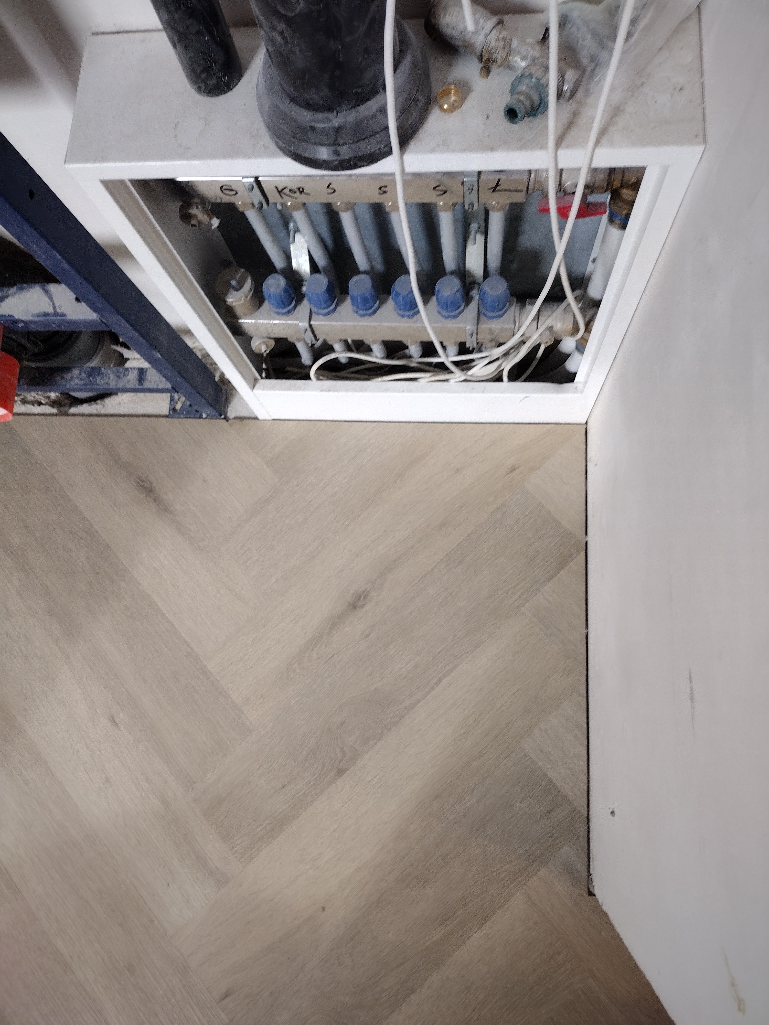 Jasny parkiet ułożony we wzór jodełki francuskiej, widoczny fragment instalacji ogrzewania podłogowego z rozdzielaczem w białej obudowie.