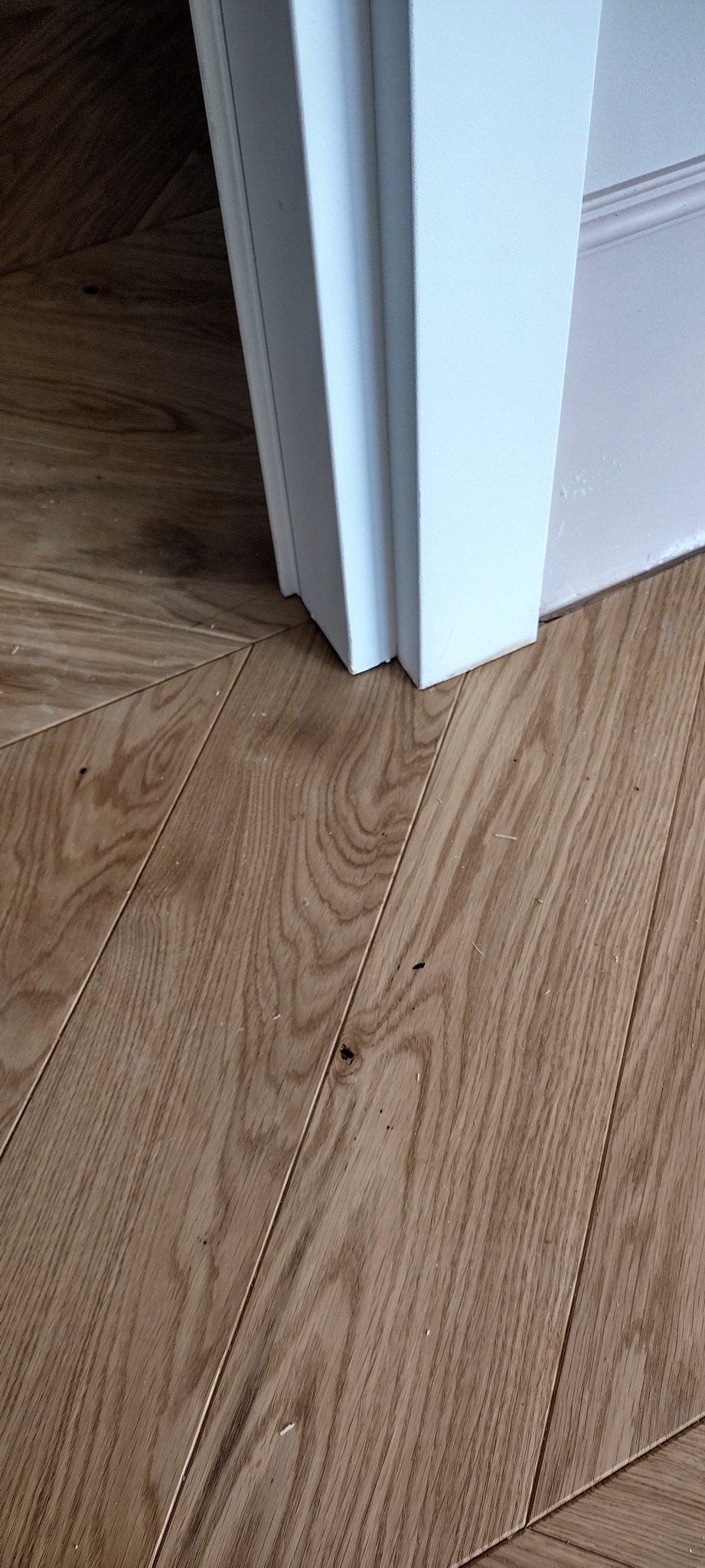 Jasnobrązowy parkiet ułożony w jodełkę, przylegający do białej ościeżnicy drzwiowej.