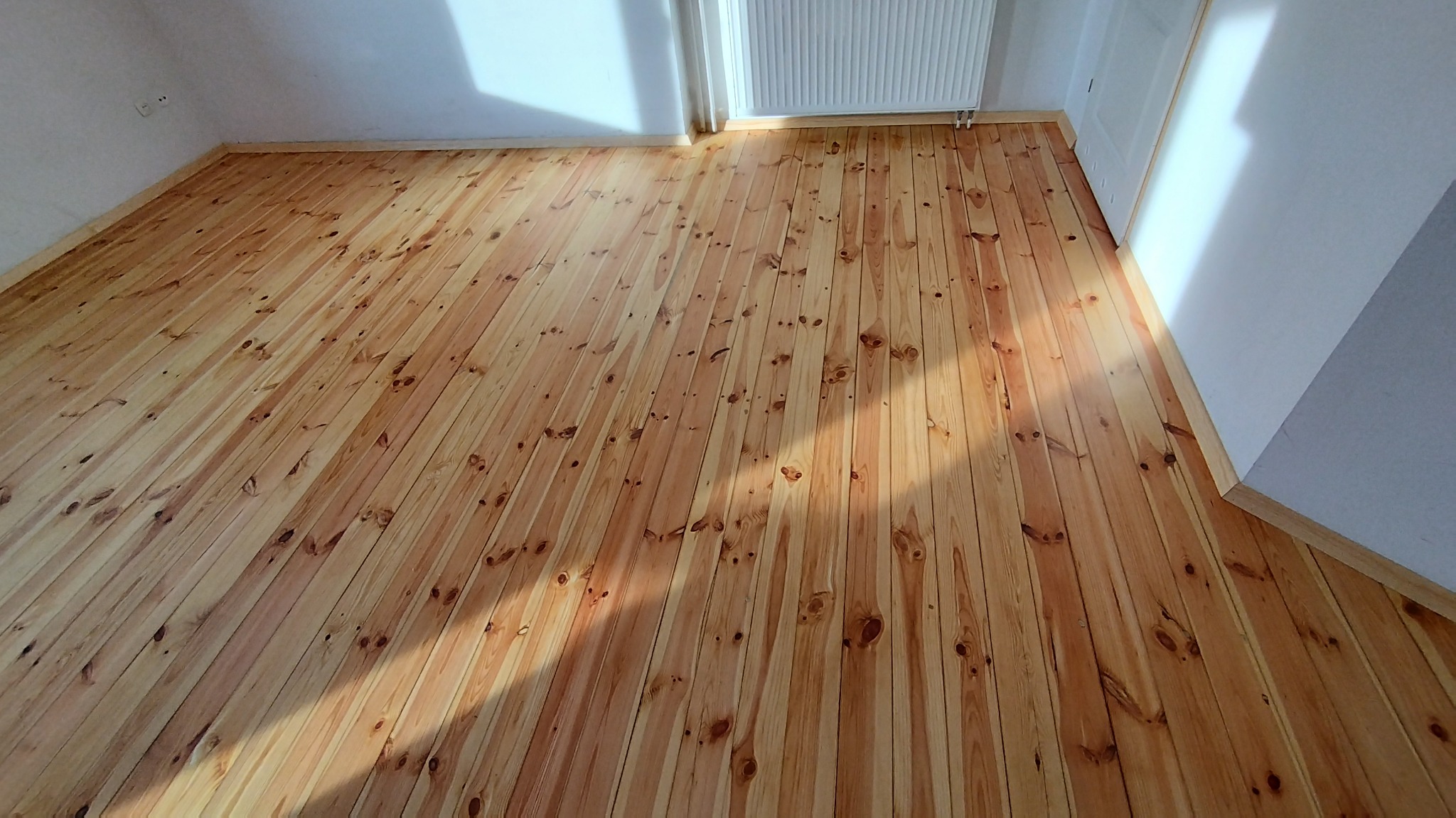 Jasny, wykończony parkiet z drewna sosnowego w pokoju z białymi ścianami, widoczne listwy przypodłogowe i grzejnik, oświetlony promieniami słońca.