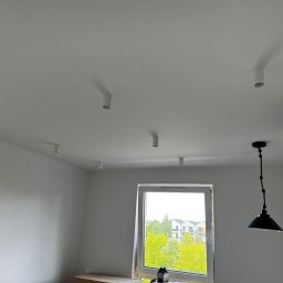 Tech Tor Patryk Christke - Wnętrze pokoju z białymi ścianami i sufitem, na którym zamontowano sześć cylindrycznych lamp sufitowych oraz jedną lampę wiszącą z czarnym kloszem w stylu industrialnym, widok na zieleń za oknem.