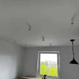 Wnętrze pokoju z białymi ścianami i sufitem, na którym zamontowano sześć cylindrycznych lamp sufitowych oraz jedną lampę wiszącą z czarnym kloszem w stylu industrialnym, widok na zieleń za oknem.