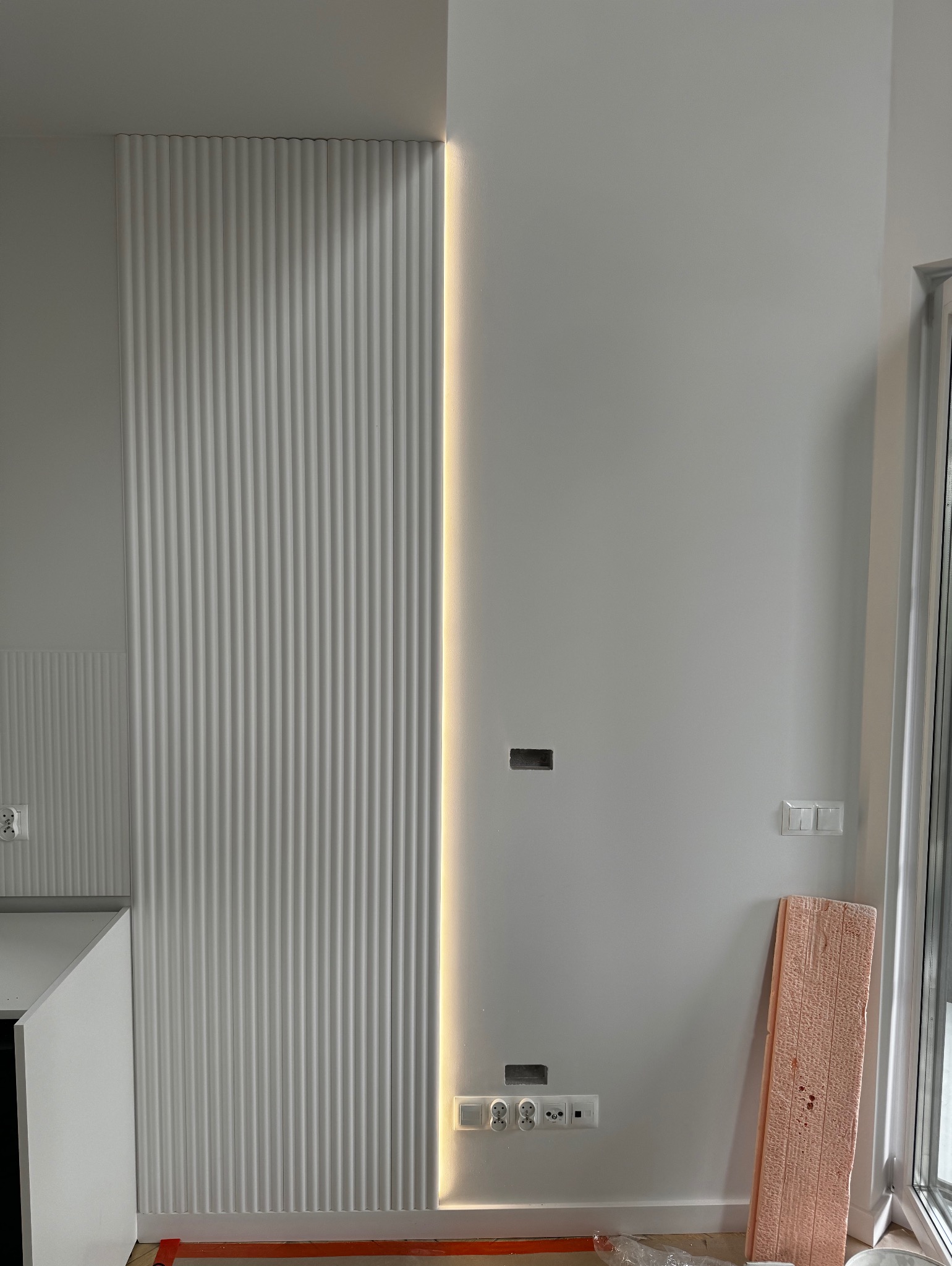 Nowoczesna ściana z dekoracyjnymi panelami pionowymi podświetlonymi paskiem LED, widoczne gniazdka elektryczne i puszki instalacyjne przed montażem osprzętu.