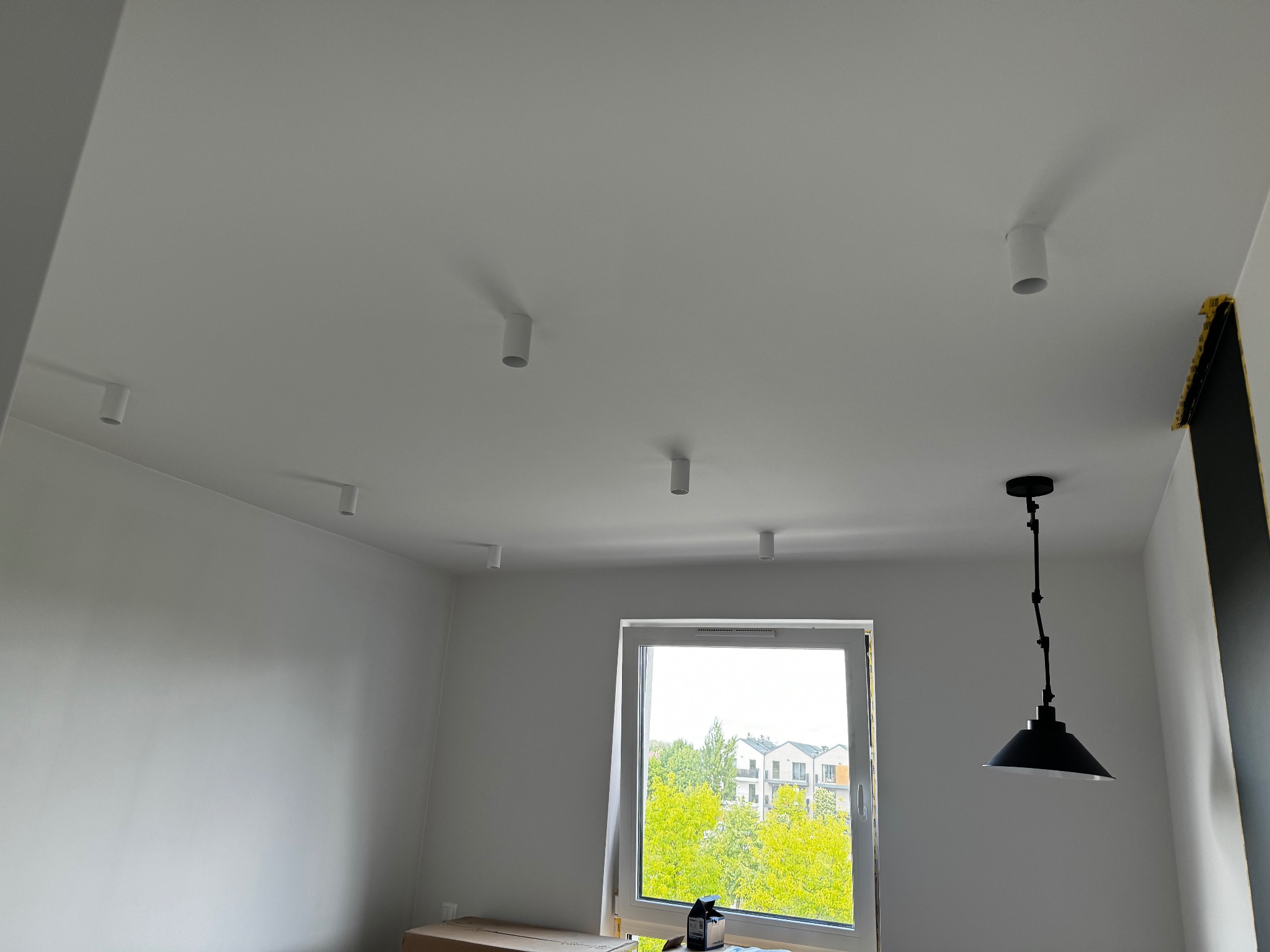 Wnętrze pokoju z białymi ścianami i sufitem, na którym zamontowano sześć cylindrycznych lamp sufitowych oraz jedną lampę wiszącą z czarnym kloszem w stylu industrialnym, widok na zieleń za oknem.