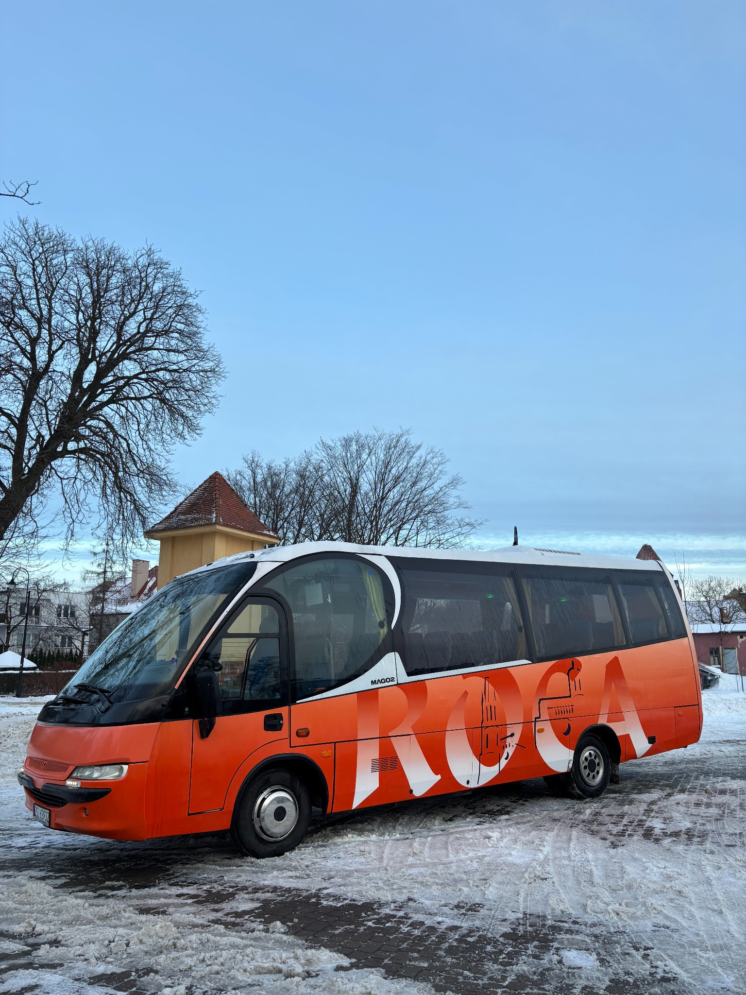Pomarańczowy bus z białym napisem ROZA na tle zimowego krajobrazu. Widoczne drzewa bez liści i budynek w oddali. Bus stoi na śniegu.