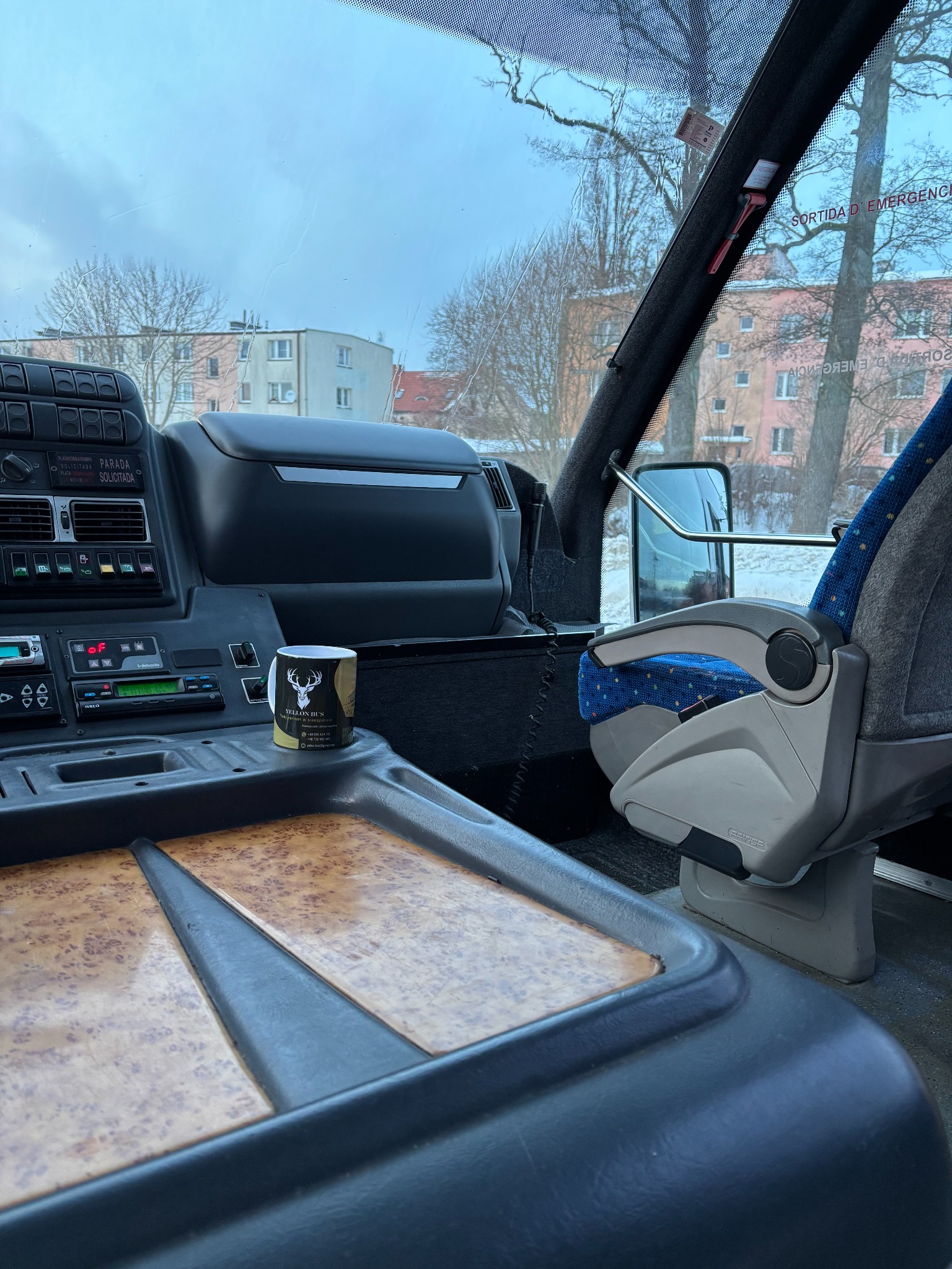 Wnętrze autobusu z widokiem na deskę rozdzielczą, fotel kierowcy i okno z budynkami w tle. Na desce stoi kubek z logo firmy transportowej.