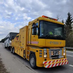 Żółty pojazd pomocy drogowej marki Renault MBT holuje biały autobus na poboczu drogi, widoczne oznaczenia firmy i numery telefonów na pojeździe, niebo pochmurne.