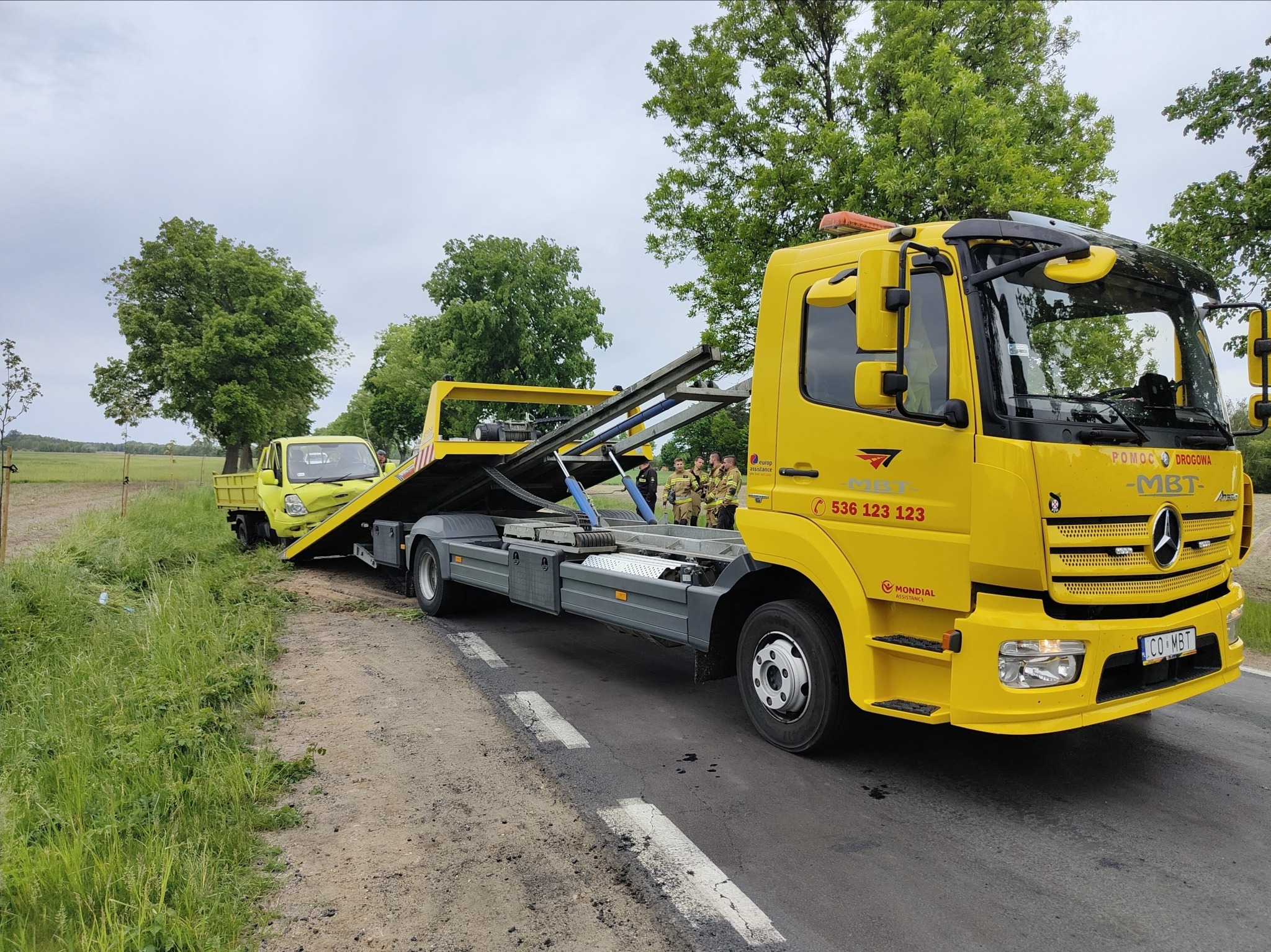 Żółta laweta z uszkodzonym, żółtym pojazdem na platformie w Sierpcu, w tle grupa osób w mundurach służb ratunkowych.