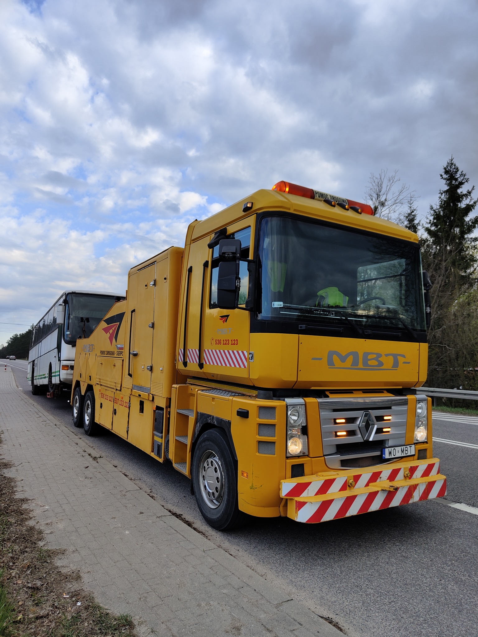 Żółty pojazd pomocy drogowej marki Renault MBT holuje biały autobus na poboczu drogi, widoczne oznaczenia firmy i numery telefonów na pojeździe, niebo pochmurne.