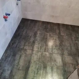 Świeżo położona, ciemna, błyszcząca glazura podłogowa w łazience z widocznymi przyłączami wodnymi na jasnych, wykafelkowanych ścianach i niedokończonym fragmentem ściany.