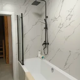 Nowoczesna łazienka z wanną obudowaną płytkami imitującymi marmur, czarną armaturą natryskową i szklanym parawanem, widok na fragment schodów w tle.