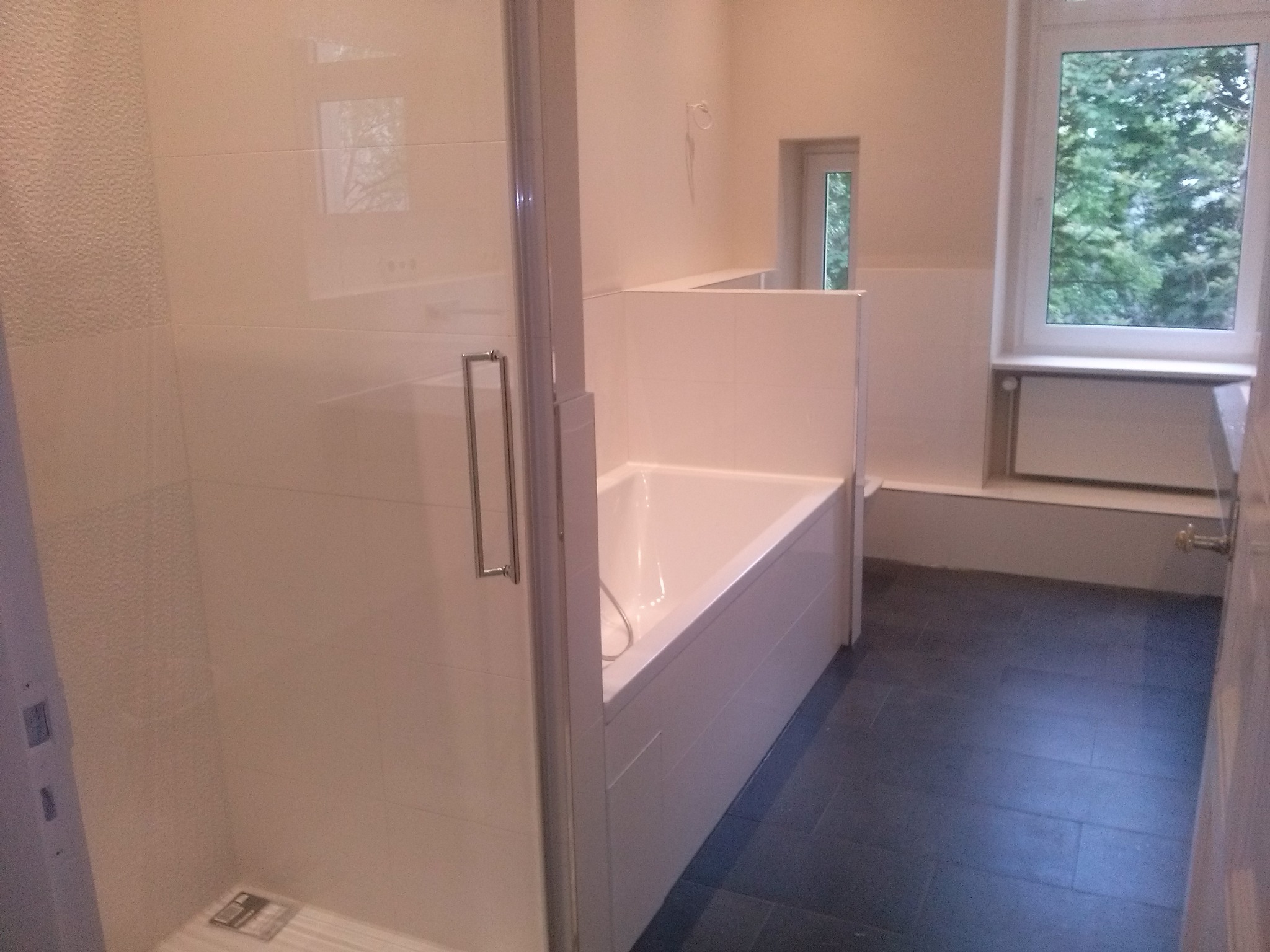 Jasna łazienka z kabiną prysznicową, wanną obudowaną płytkami i ciemną podłogą z płytek, widok z perspektywy drzwi wejściowych.