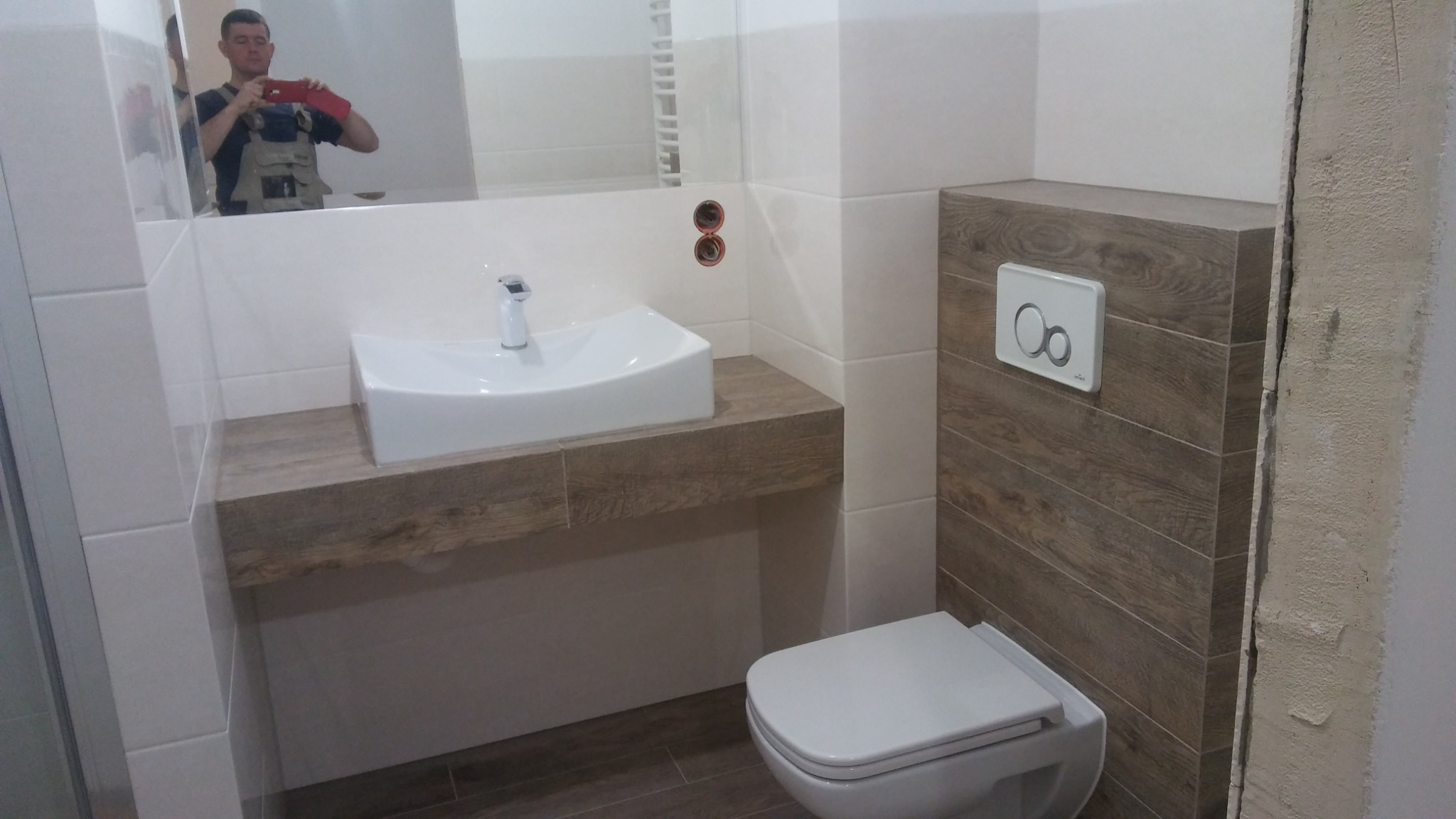 Nowoczesna łazienka z białą umywalką nablatową na drewnopodobnym blacie, wisząca toaleta z drewnopodobną obudową i białym przyciskiem spłukującym. W lustrze odbija się osoba robiąca zdjęcie telefonem.