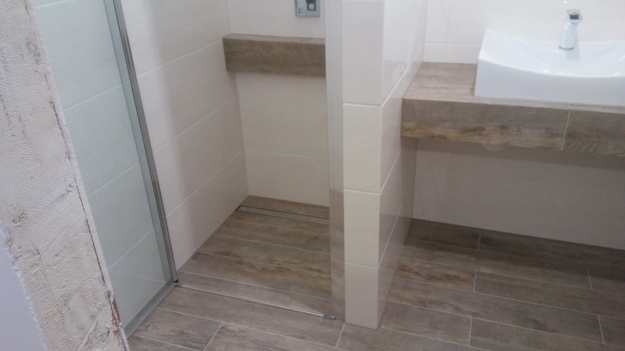 Nowoczesna łazienka z kabiną prysznicową z szklanymi drzwiami, umywalką nablatową na blacie z płytek imitujących drewno i odpływem liniowym w podłodze z płytek drewnopodobnych.