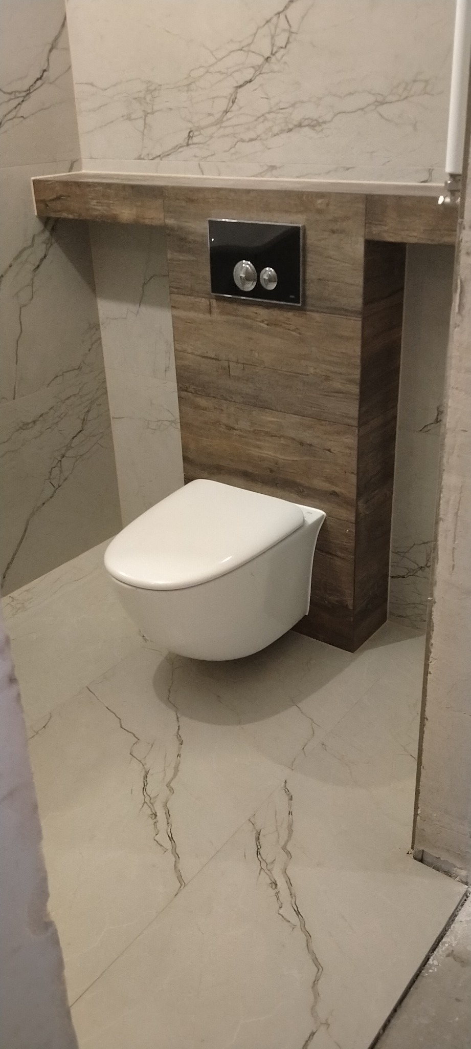 Wisząca toaleta z podtynkową spłuczką na tle marmurowych płytek ściennych i podłogowych, drewnopodobna obudowa stelaża.