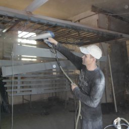 Diuneto - Pracownik w czapce maluje metalowe elementy metodą natryskową w warsztacie, używając pistoletu lakierniczego.