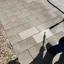 Czyszczenie kostki brukowej myjką ciśnieniową, widoczna różnica w kolorze między umytą a brudną powierzchnią, zielony wąż i osoba w zielonej koszuli w kratę.