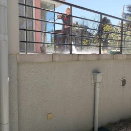 Diuneto - Pracownik w kombinezonie roboczym czyści balkon z czarną balustradą za pomocą myjki ciśnieniowej, w tle widoczne drzewa i budynki.