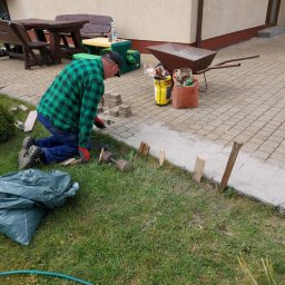 Diuneto - Pracownik w zielonej koszuli w kratę układa kostkę brukową na posesji, widoczny młotek, worki z materiałami, taczka z piaskiem i wytyczone drewniane kołki.