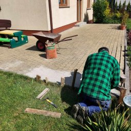 Diuneto - Mężczyzna w zielonej koszuli w kratę klęczy na trawie, układając kostkę brukową na podwórku. Widoczna taczka, worek z materiałem, młotek i drewniane elementy szalunku.