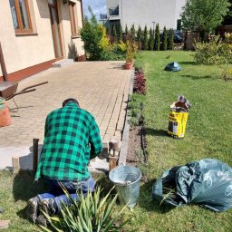 Diuneto - Mężczyzna w zielonej koszuli w kratę klęczy przy krawężniku z kostki brukowej, przygotowując rabatę na lawendę, obok stoi wiadro i worek z ziemią.