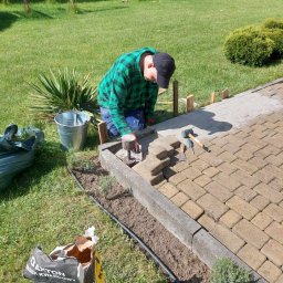 Diuneto - Układanie kostki brukowej na trawniku w ogrodzie, widoczny pracownik w kraciastej koszuli, narzędzia, worek z piaskiem kwarcowym Axton, częściowo ułożony chodnik, elementy drewniane jako ograniczniki.