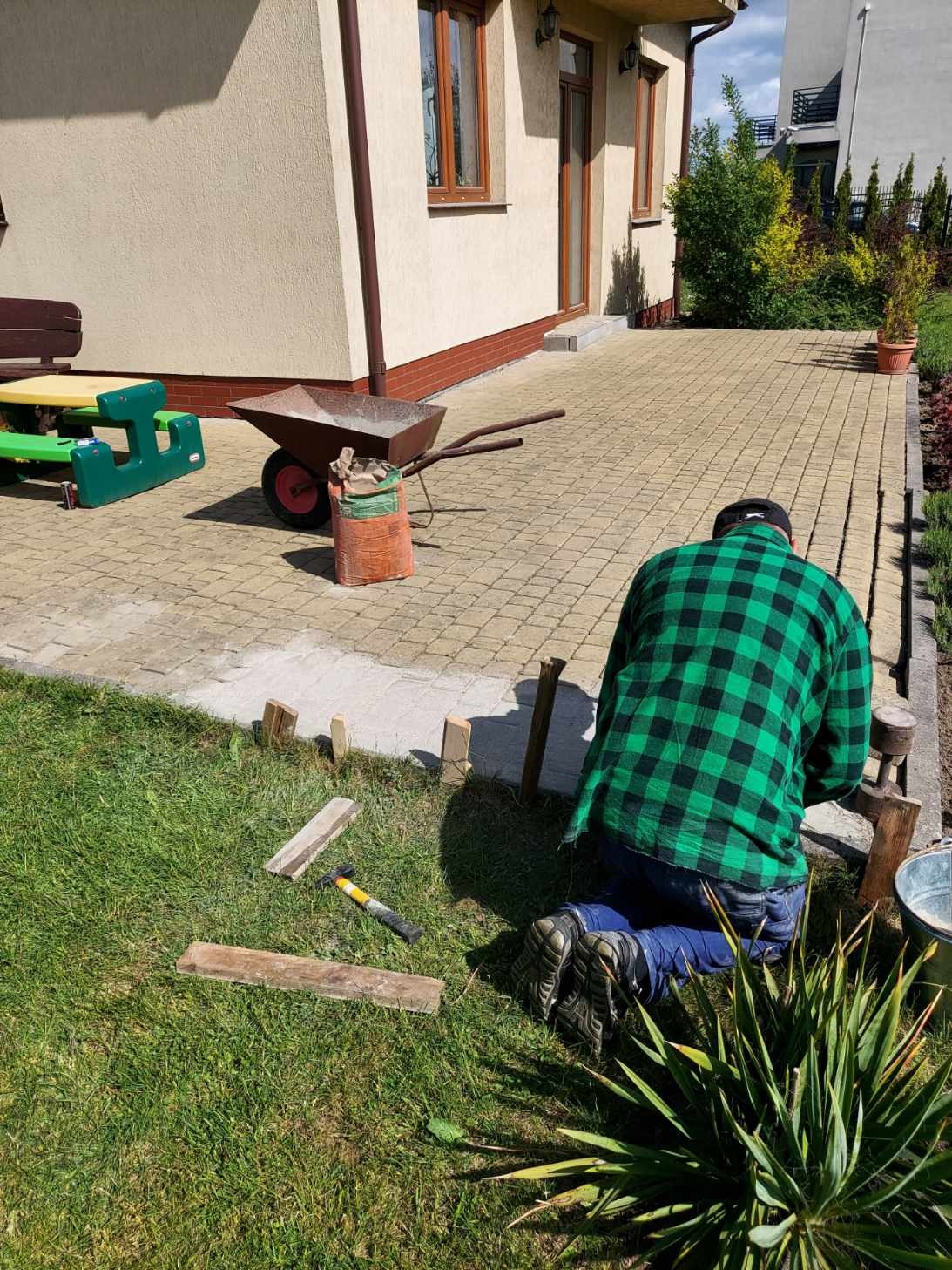 Mężczyzna w zielonej koszuli w kratę klęczy na trawie, układając kostkę brukową na podwórku. Widoczna taczka, worek z materiałem, młotek i drewniane elementy szalunku.
