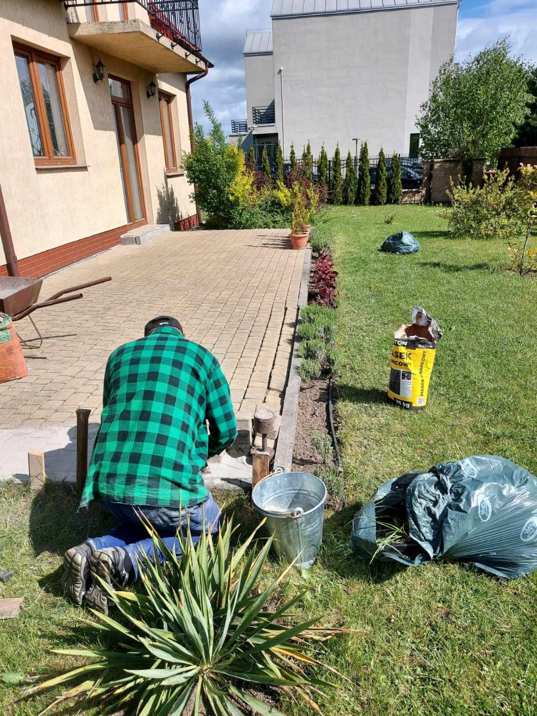 Mężczyzna w zielonej koszuli w kratę klęczy przy krawężniku z kostki brukowej, przygotowując rabatę na lawendę, obok stoi wiadro i worek z ziemią.