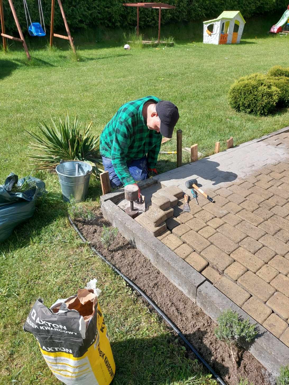 Układanie kostki brukowej na trawniku w ogrodzie, widoczny pracownik w kraciastej koszuli, narzędzia, worek z piaskiem kwarcowym Axton, częściowo ułożony chodnik, elementy drewniane jako ograniczniki.