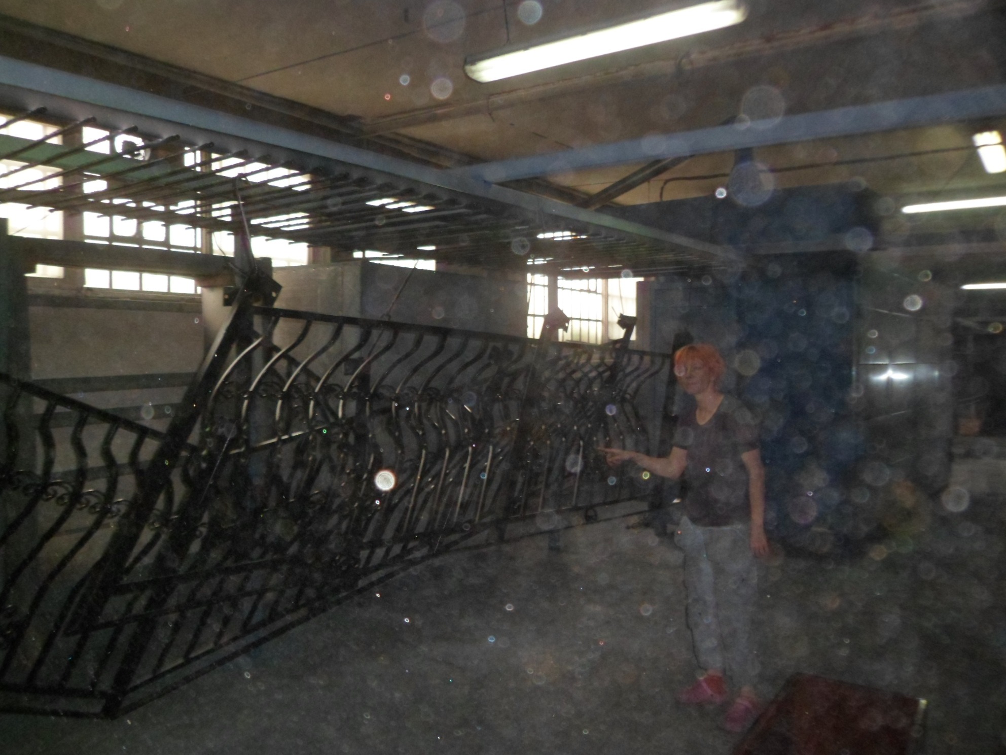 Część metalowego ogrodzenia z ozdobnymi elementami, prezentowana przez osobę w hali produkcyjnej z widocznymi oknami w tle.