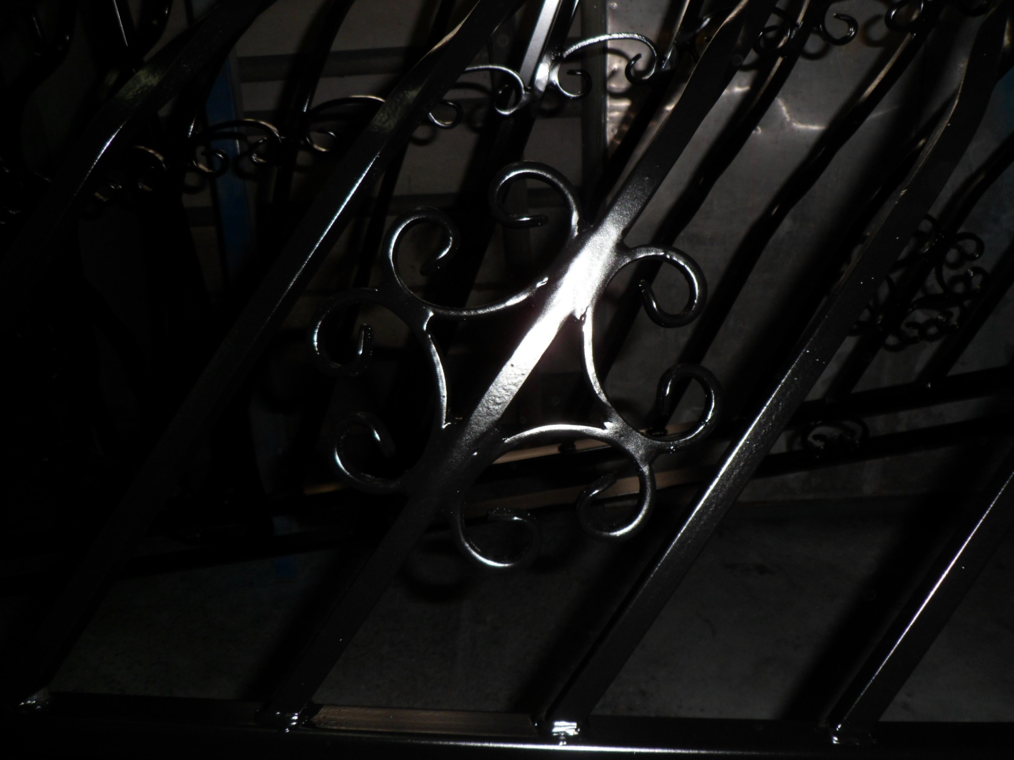 Metalowa brama z dekoracyjnymi elementami w kształcie spiral, oświetlona punktowo, widoczna z bliska w ciemnym otoczeniu.