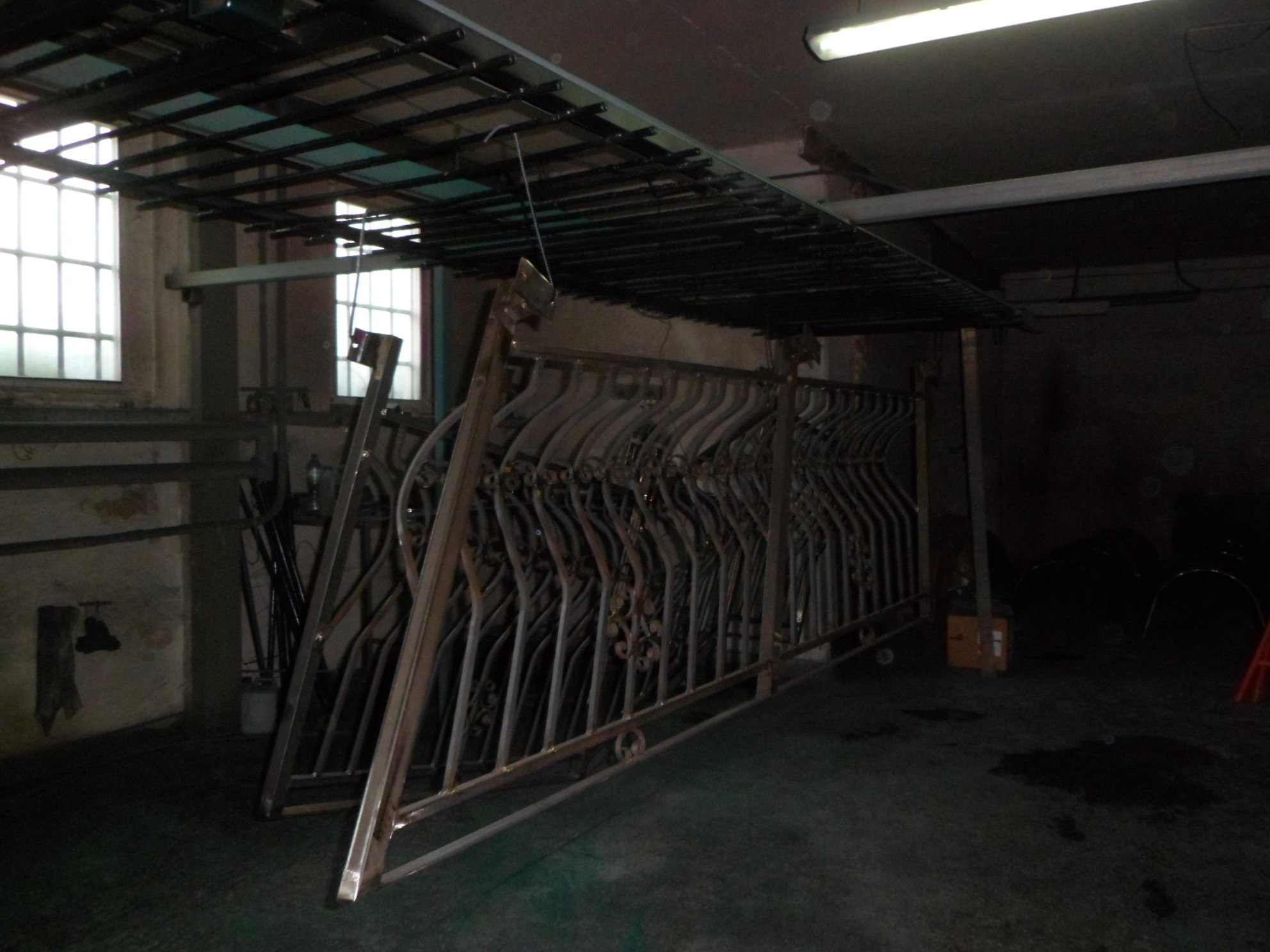 Stos metalowych, ozdobnych balustrad ustawionych w warsztacie pod metalową konstrukcją, oświetlonych jarzeniówką.