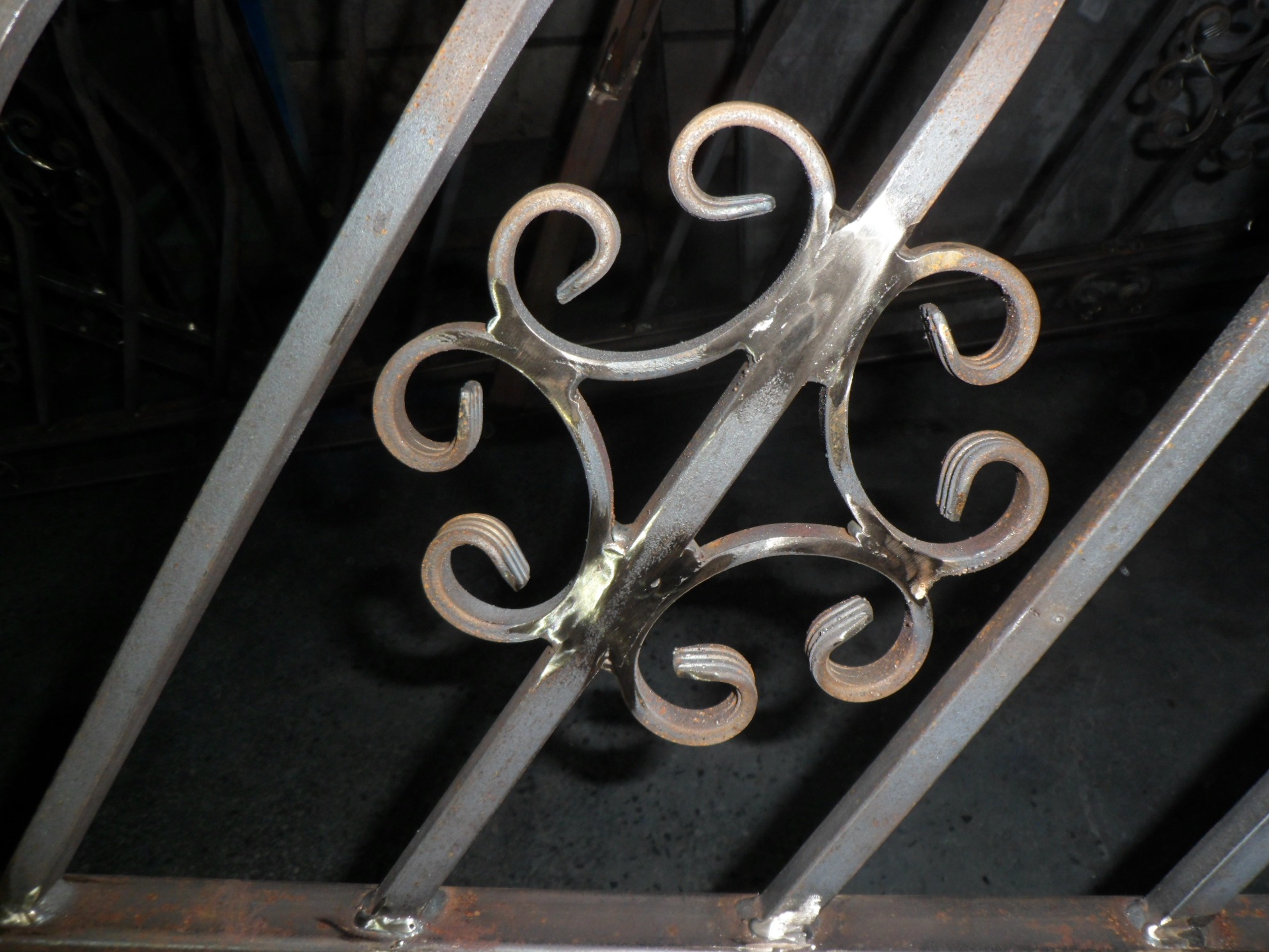 Metalowa krata z ozdobnym, spiralnym elementem centralnym, surowe wykończenie, widoczne ślady spawania.