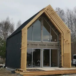 Surowy stan domu w trakcie budowy, widoczna konstrukcja szkieletowa z drewna, czarna blacha na elewacji, duże okna z widocznym wnętrzem i drabina w środku, teren budowy z gruzem i materiałami.