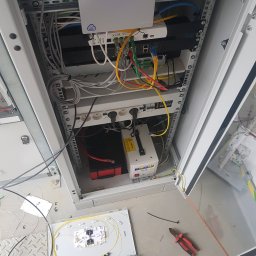 P.H.U ANDRZEJ GRZESIAK - Otwarta szafa rack z zamontowanymi urządzeniami sieciowymi, zasilaczem awaryjnym i patch panelem światłowodowym na podłodze, obok leżą obcęgi.